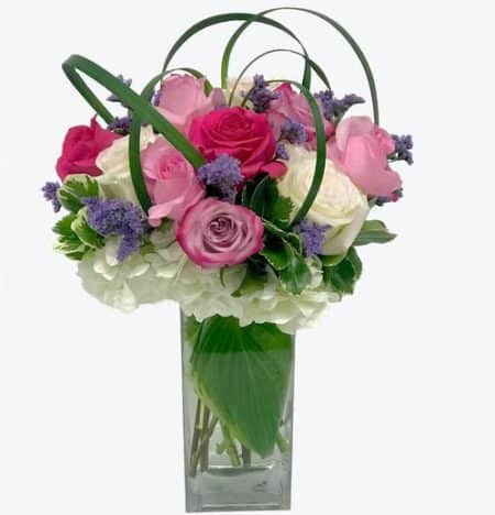 A modern compact design of pastel roses in lavender, pinks and whites nestled in a hydrangea with dancing lily grass loops. Designed in a glass vase. (vase may vary)